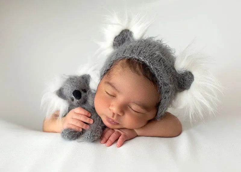 A newborn baby sleeps peacefully wearing a grey bonnet against a white backdrop.