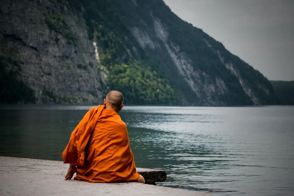 A monk wearing  orange cloth sitting next to a lake