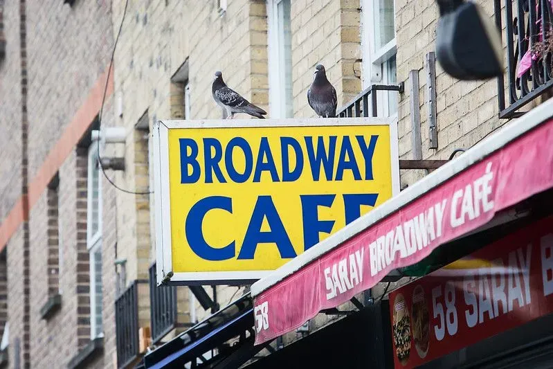 A yellow sign at Broadway Market reading 'Broadway Cafe' with pigeons sitting on it.