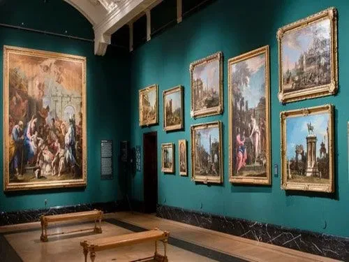A teal wall at The Queen's Gallery covered with paintings in gold frames.