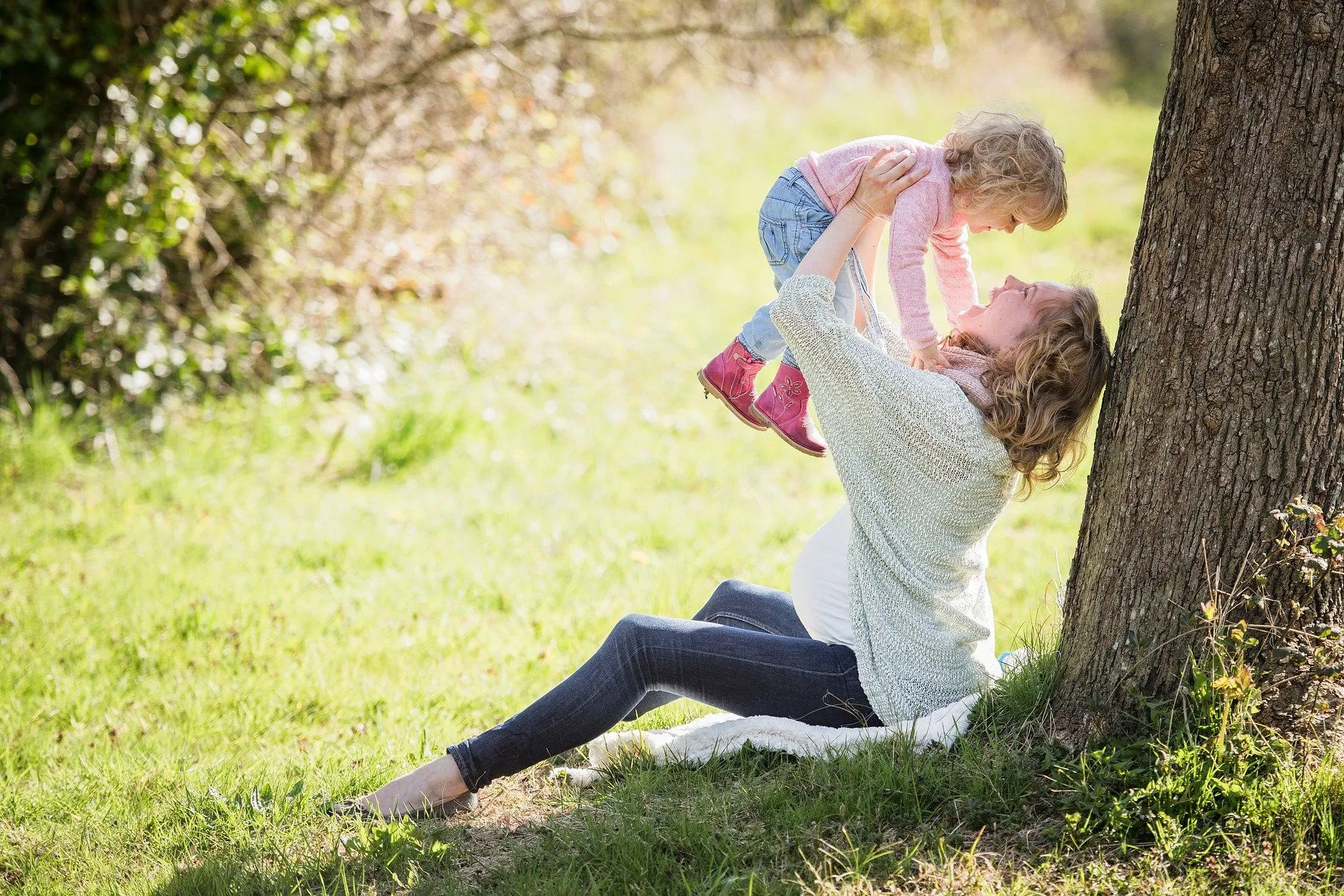 A mother raises her small daughter into the air, while sat beneath a tree in summer time.