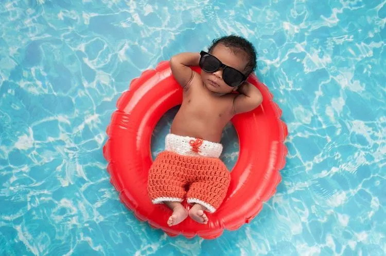 Two week old newborn baby boy sleeping on a tiny inflatable swim ring wearing crocheted board shorts and black sunglasses.