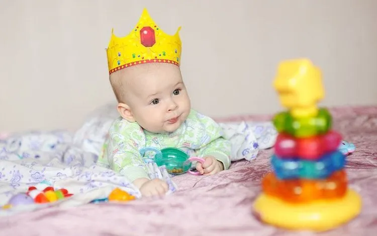 A newborn baby wearing a crown lies on a bed with toys