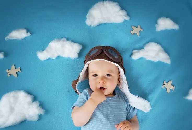 A newborn baby boy wearing vintage pilot's hat and lying next to clouds and airplanes