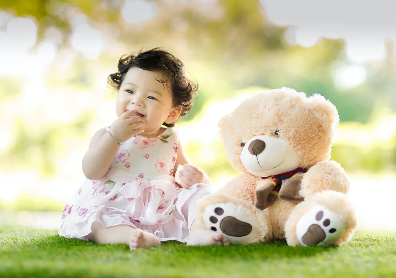Baby sitting on green grass beside bear plush toy at daytime.
