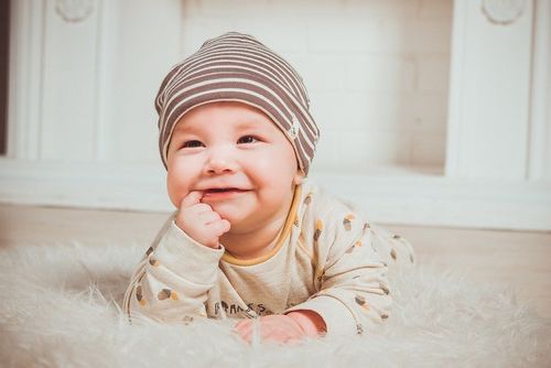 Smiling baby wearing a hat lying on stomach and its chewing hands.