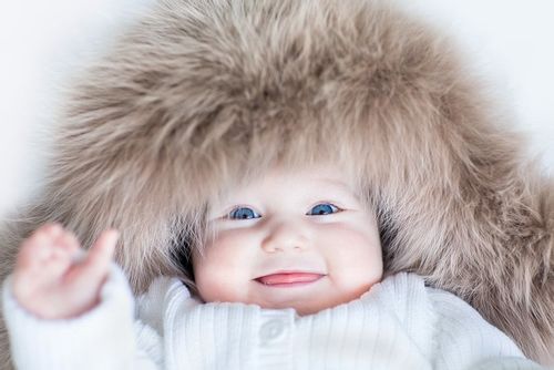 Blue-eyed baby lying on its back wearing a furry hood smiling.