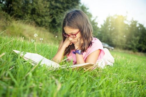 Young girl lying on her stomach in the grass reading a book of Victorian poetry.