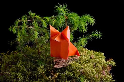A red origami wolf placed on artificial grass with artificial trees behind.