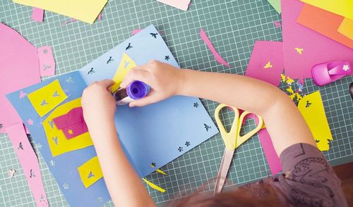 Little girl sat at a table making an origami birthday card, sticking on designs with glue.