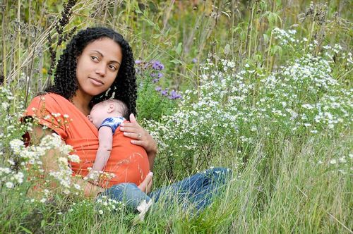 Mum sat on the floor in a field with her child in a baby sling and flowers around them.