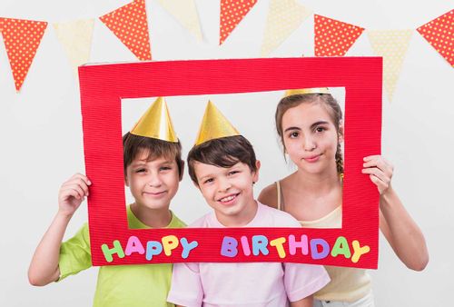 Three kids holding up a birthday banner frame and standing with their faces inside.