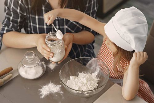 Young girl in a chef's hat adding a spoon of flour into her cake mix from the jar that her mum is holding for her.