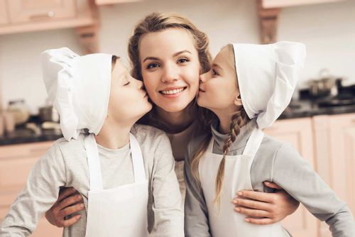Two children, wearing chef's hats, kissing their mum on each cheek as they bake in the kitchen.