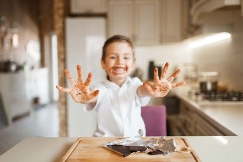 Little girl in the kitchen holding her hands out in front of her showing the chocolate on them and around her mouth too.