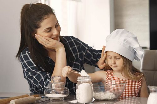 Mother smiles at daughter wearing a chef hat as she weighs flour to make a cow cake.