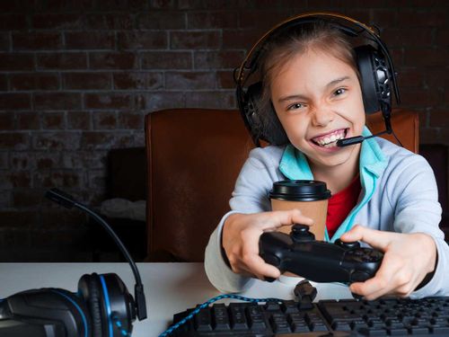 A young girl wearing headphones and holding a remote control smiles at the camera as she plays an Xbox game.
