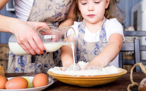 A little girl mixes flour and milk in a bowl with her hands to make a snake cake whilst her mother pours more milk into the bowl from above.