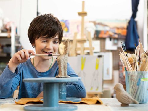 A boy smiles as he uses a craft tool to decorate a giraffe clay model.