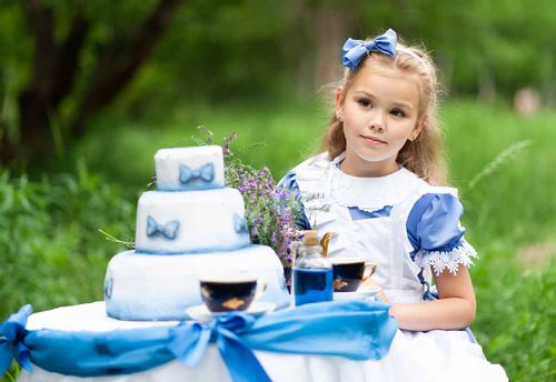 Little girl dressed as Alice in Wonderland sat at a table outside having a tea party with a tiered blue and white cake.
