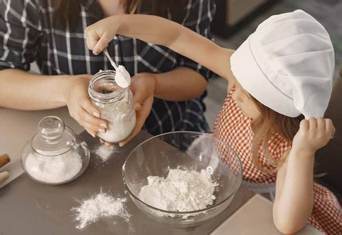 Little girl in a chef's hat spooning flour into a large mixing bowl to make the batter of a frog cake.