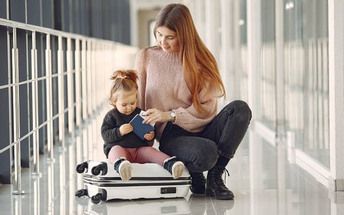 Mum crouching next to her young toddler who is sat on the suitcase at the airport.