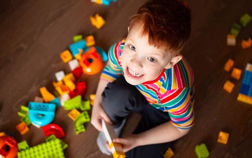Little boy crouching on the floor playing with his Lego, smiling up at the camera.