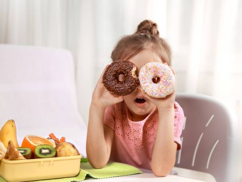 Little girl sat at the table holding up two doughnuts in front of her eyes making a silly face.