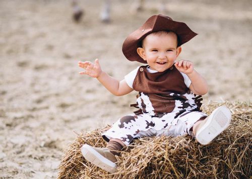 Baby boy dressed as a cowboy, smiling as he sits on a bale of hay.