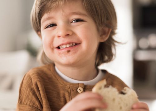 Little boy holding a sandwich in his hand as he eats it, smiling, with the brown filling on his face.