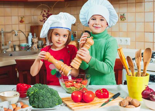 Two kids standing in the kitchen wearing chef's hats as they season a salad in the bowl.