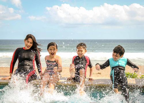 A family wearing wetsuits smile as they splash sea water with their feet at the beach.
