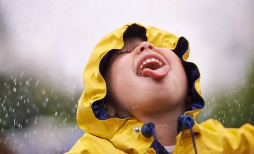Little child wearing a yellow raincoat, hood up, standing in the rain and sticking out their tongue to catch the falling raindrops.
