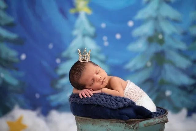 A newborn baby sleeping on a blue pillow wearing a tiny crown