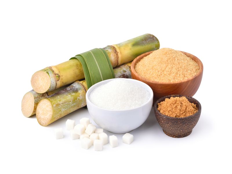 Different types of sugar are in a white bowl and a wooden bowl with fresh sugarcane.